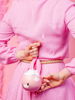 A woman dressed head to toe in pink strikes a match over a pink Cloud stoneware mug printed with a black cat, a candle burning inside it