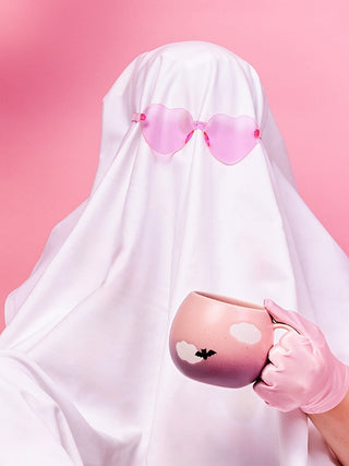 A ghost in a white sheet with pink heart sunglasses holding the pink Cloud mug printed with a bat
