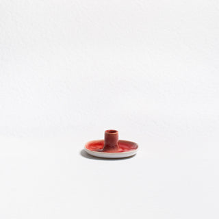 Angled view of the red Christmas candle holder — a shallow round stoneware dish with a central socket for a taper candle, red glaze pooling over a white rim.